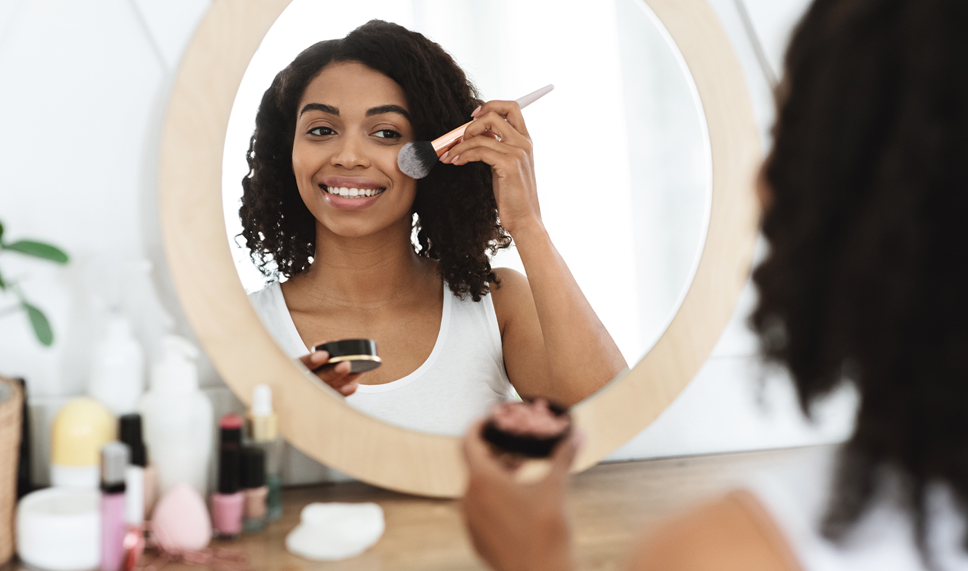 Beautiful black woman applying blush on face, using brush tool, putting makeup